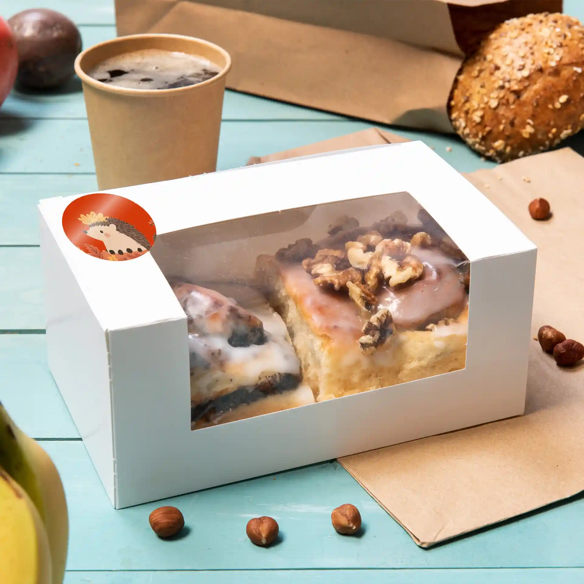 White box with a transparent window containing two pastries topped with icing and walnuts, surrounded by a coffee cup and snacks.