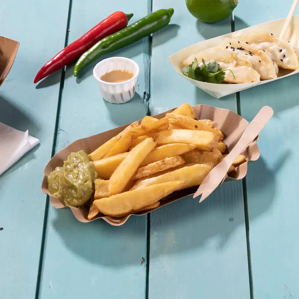 Korb mit Pommes frites, grüner Sauce, Holzutensilien und Chili auf einem blauen Holztisch.
