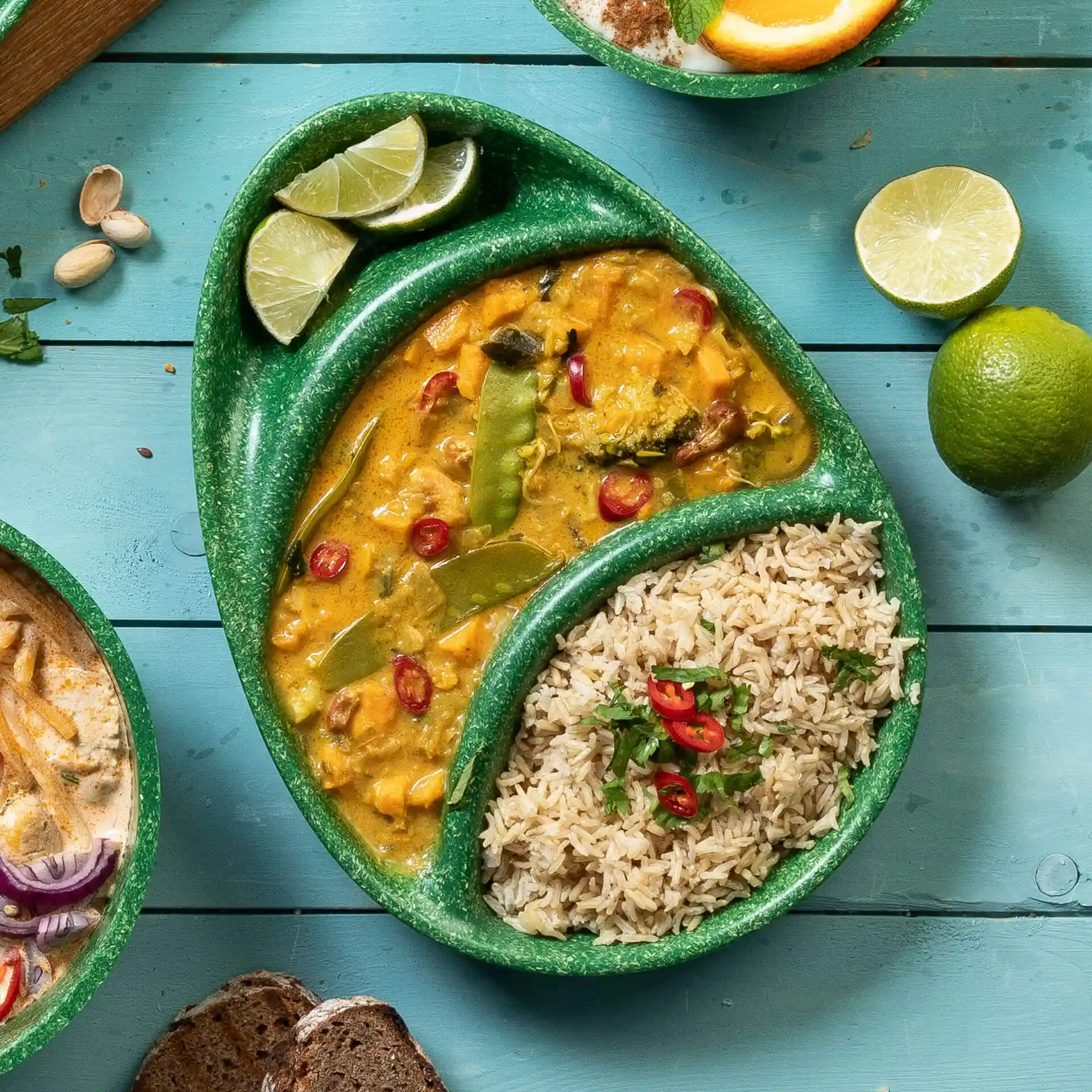 Grüner wiederverwendbarer Teller mit drei Fächern, gefüllt mit Curry, Reis und Limettenstücken auf einer blauen Holzoberfläche.