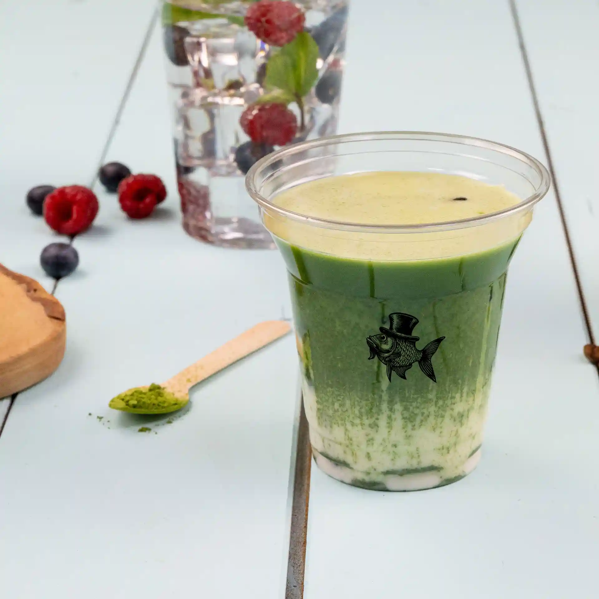 Klarer 8 oz rPET-Becher gefüllt mit grünem Getränk, mit Fisch-Logo, Holzlöffel und Beeren im Hintergrund.