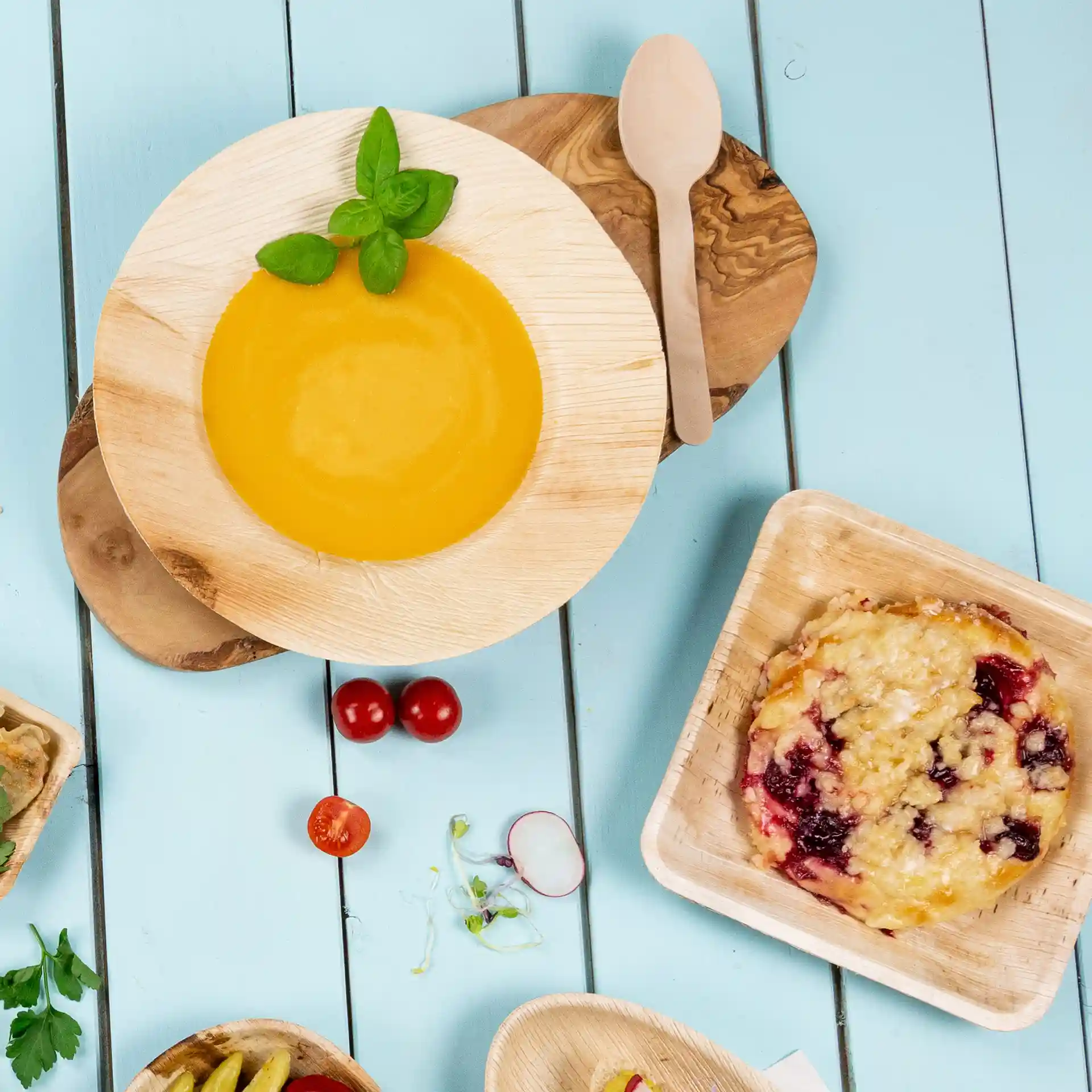Tiefe runde Teller aus Palmblatt mit gelber Suppe, garniert mit Basilikum, und einem Holzlöffel auf einer blauen Holzoberfläche.