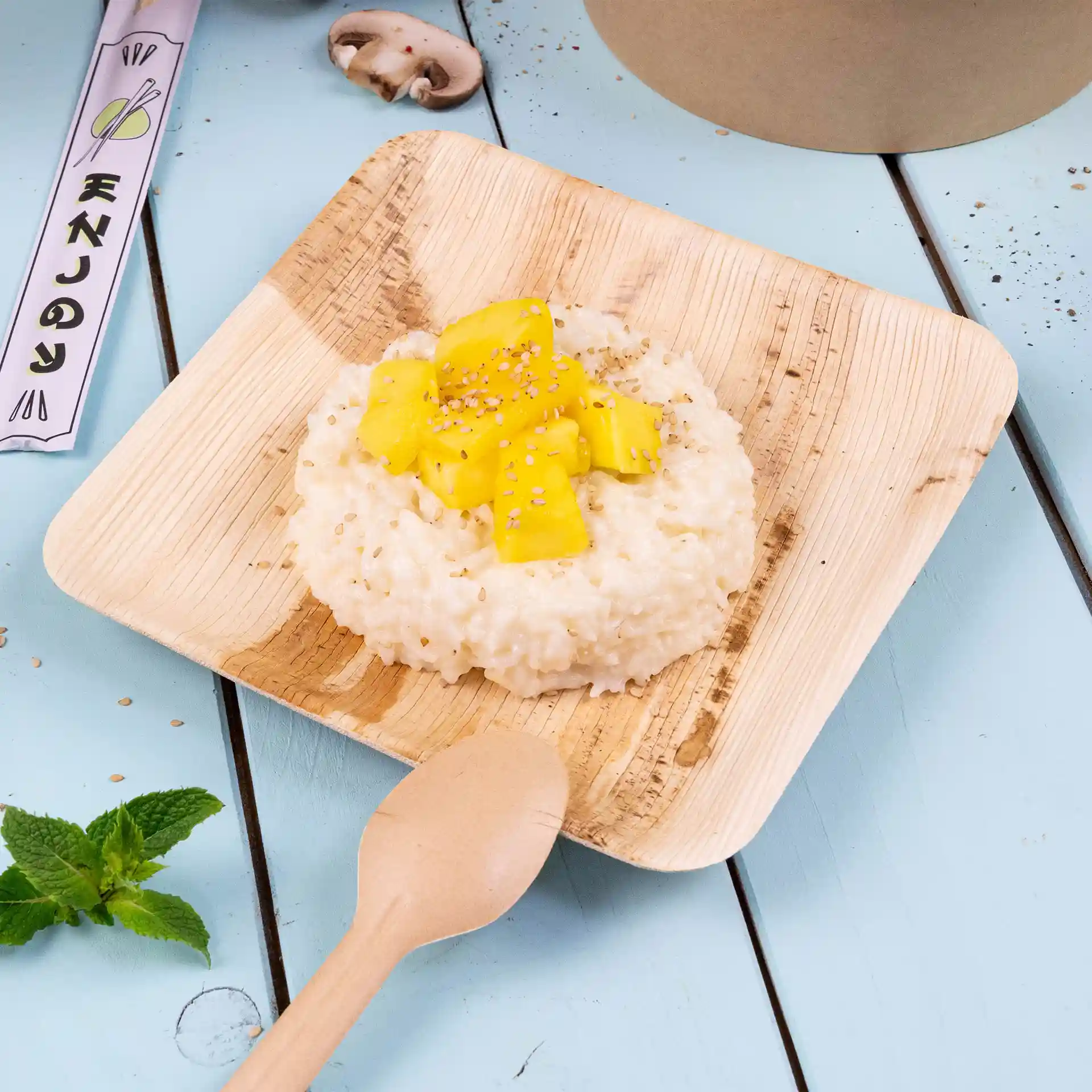 Square palm leaf plate with risotto topped with mango and sesame seeds, accompanied by a wooden spoon on a blue surface.