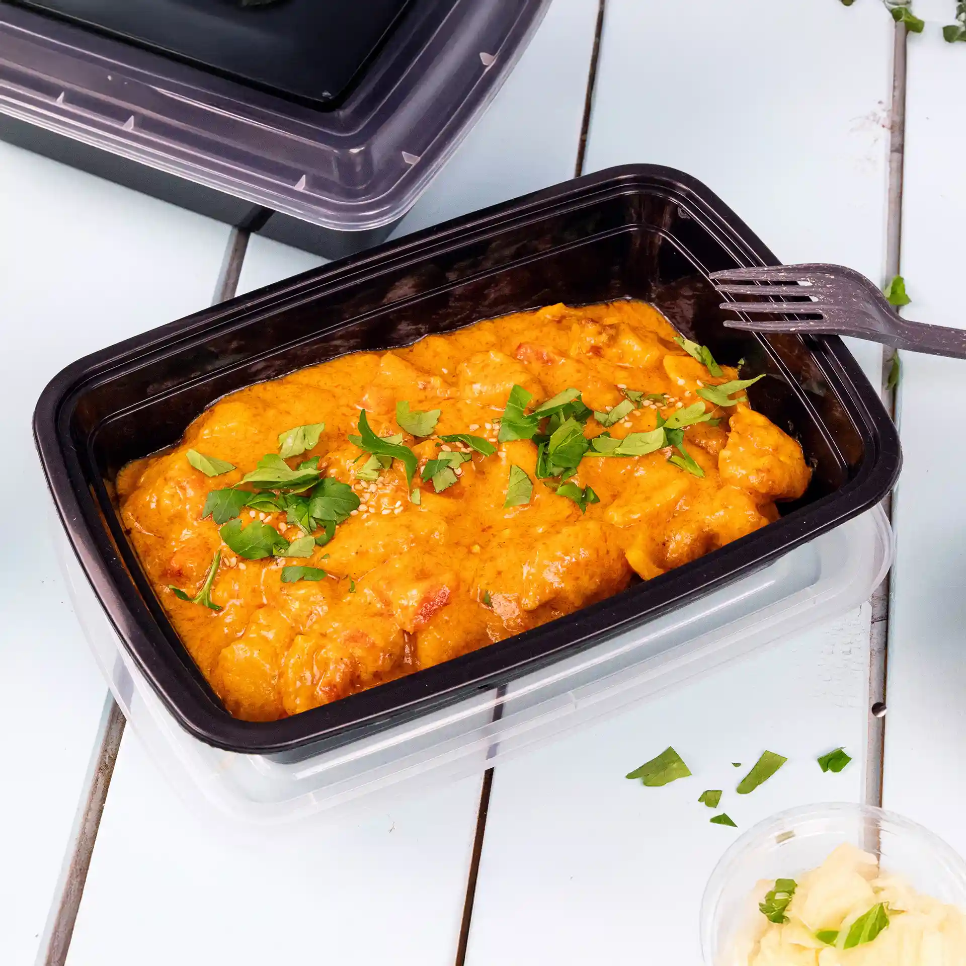 Rechteckiger schwarzer wiederverwendbarer Behälter mit Deckel, gefüllt mit orangefarbenem Essen, garniert mit grünen Kräutern und Sesam.