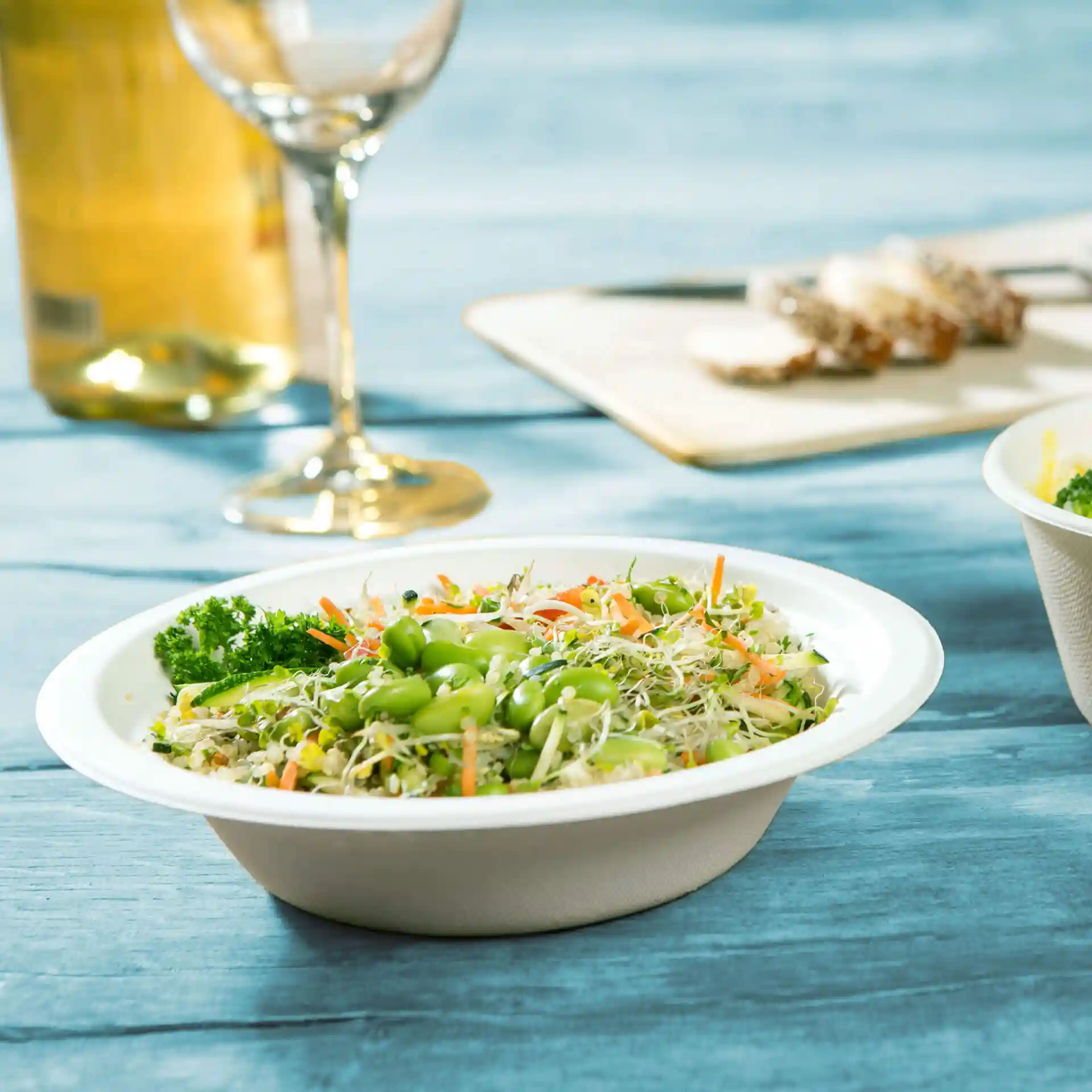Runde weiße Zuckerrüben-Schüssel gefüllt mit gemischtem Salat, einschließlich Edamame, Karotten und Sprossen, auf einer blauen Holzoberfläche.