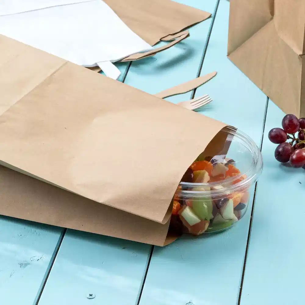 Braune Papiertüten, die teilweise einen klaren Behälter mit Salat auf einem blauen Holztisch verdecken. Trauben sind in der Nähe sichtbar.