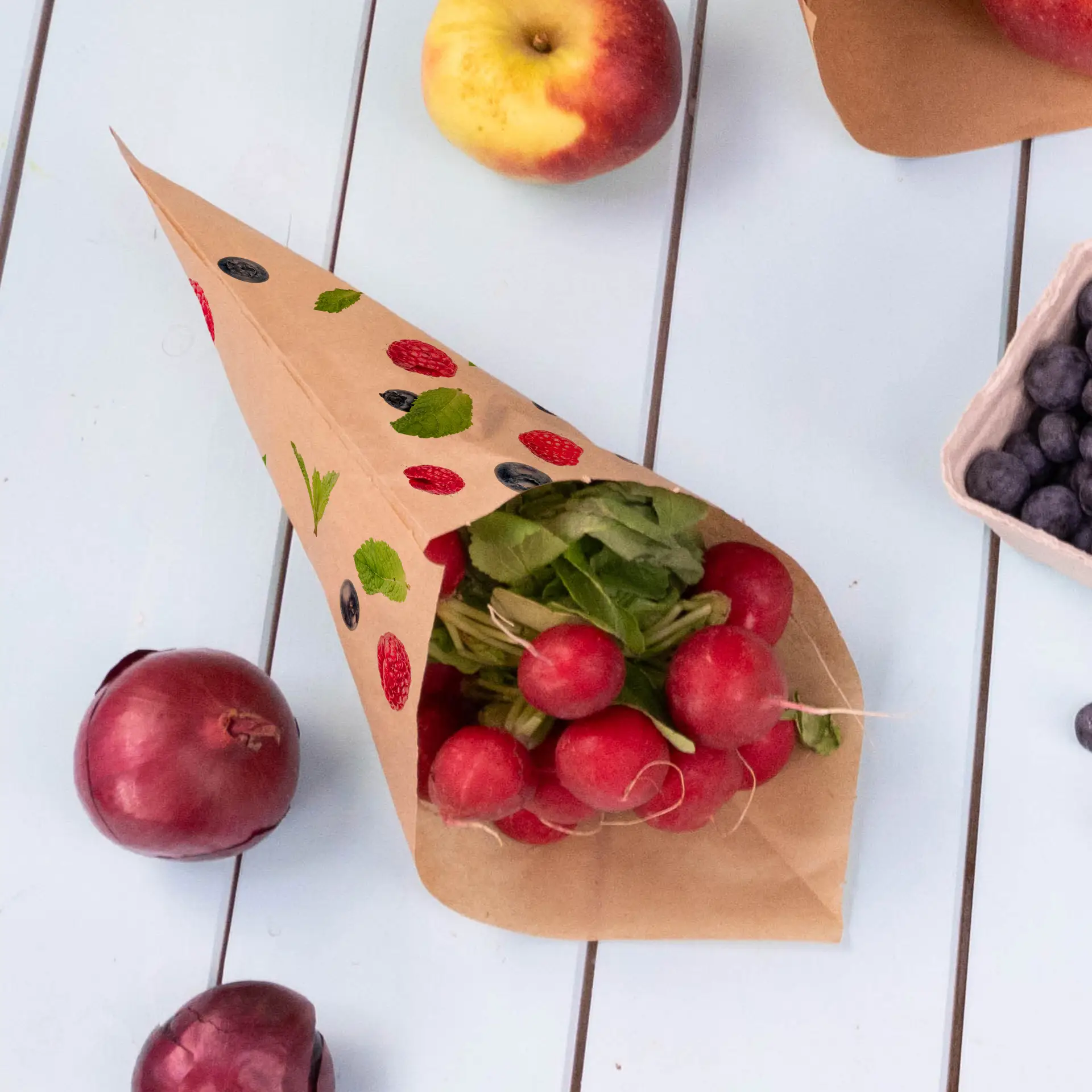 Brauner Papiertütenkonus gefüllt mit Radieschen und Kräutern, umgeben von Äpfeln, roten Zwiebeln und Heidelbeeren auf einer hellen Fläche.