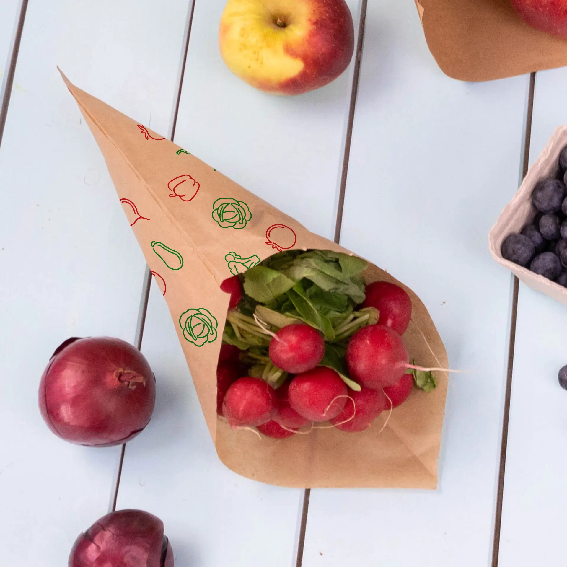 Brown paper cone bag filled with radishes and herbs, surrounded by apples, red onions, and blueberries on a light surface.