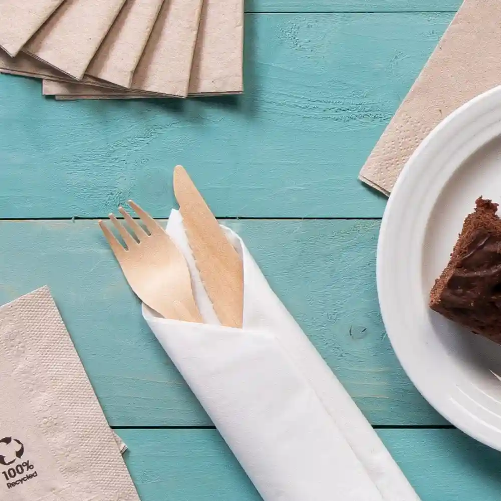 Wooden cutlery wrapped in a white napkin, accompanied by recycled paper napkins and a piece of cake on a plate.