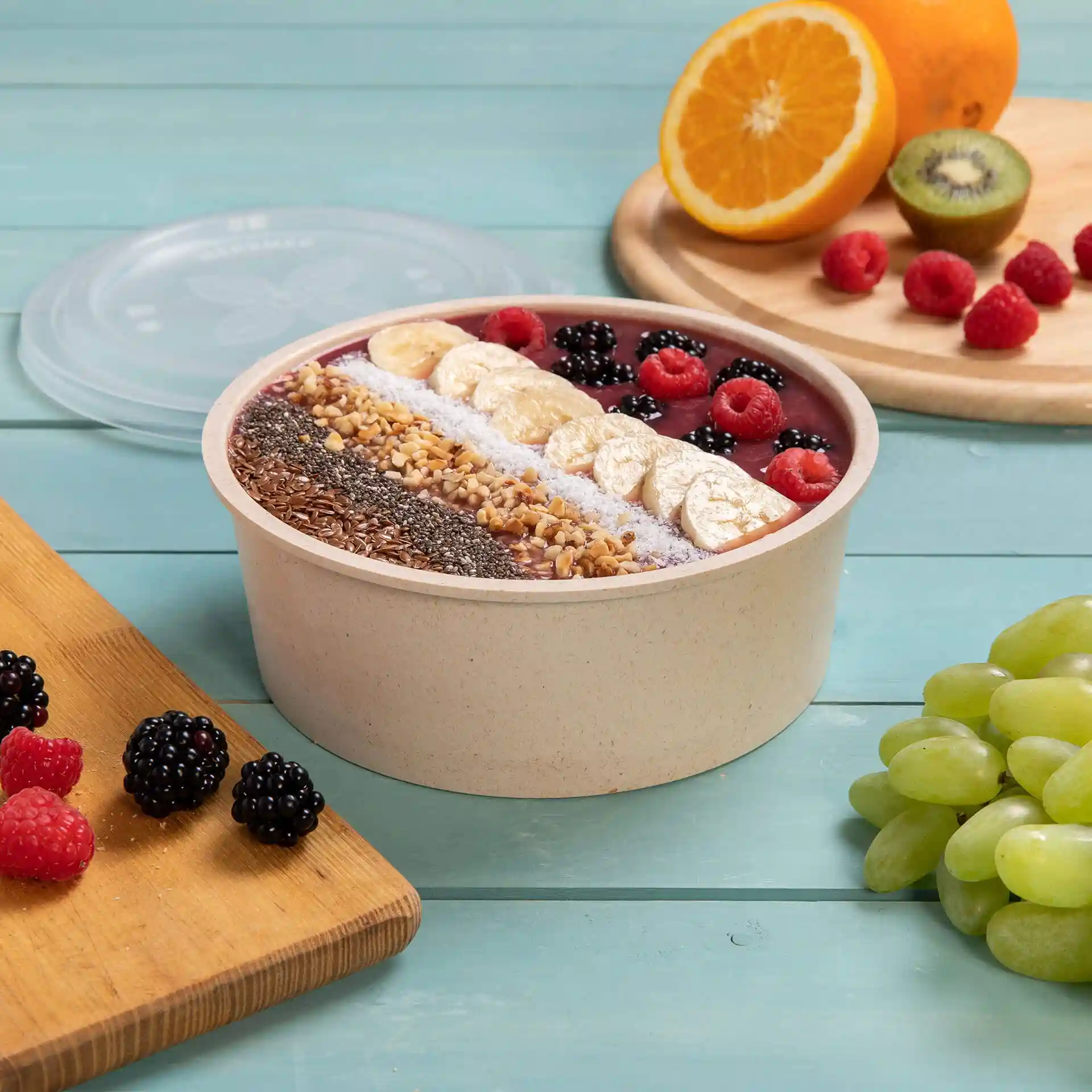 Runde wiederverwendbare Schüssel gefüllt mit Smoothie und belegt mit Früchten, auf einer blauen Holzoberfläche mit weiteren Früchten daneben.
