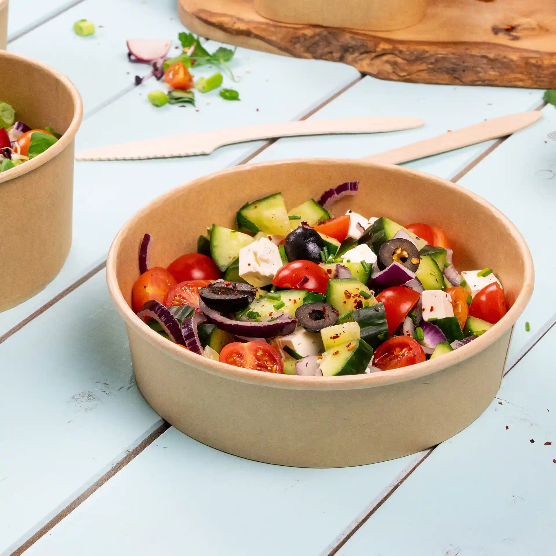 Runde braune Pappschüssel gefüllt mit einem Salat aus Tomaten, Gurken, Oliven und Feta-Käse.