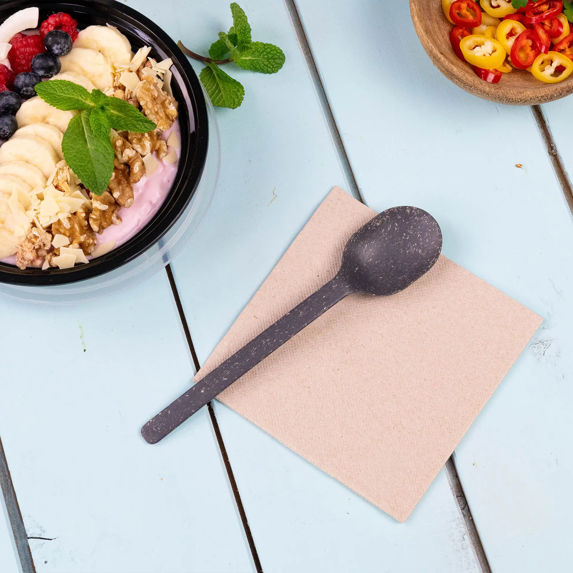 Image of a black spoon on a beige napkin, with a bowl of yogurt topped with fruit and nuts in the background.