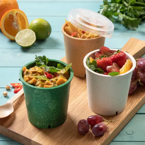 Drei wiederverwendbare Suppenbecher auf einem Holzbrett, gefüllt mit Suppe, Obst und Nudeln, umgeben von frischen Zitrusfrüchten und Trauben.