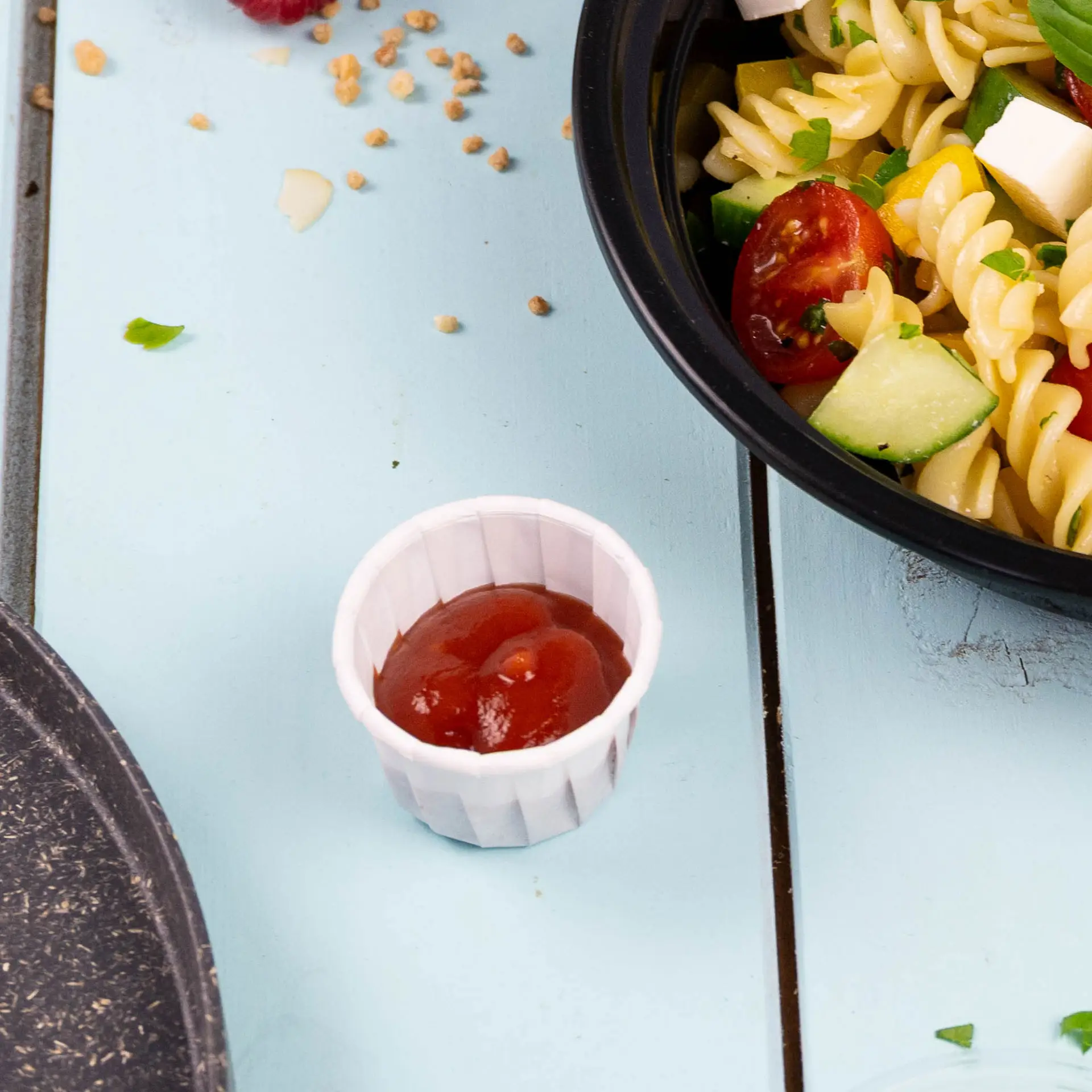 Kleiner weißer Becher mit Ketchup, neben einer Schüssel Nudelsalat auf einer blauen Holzoberfläche.