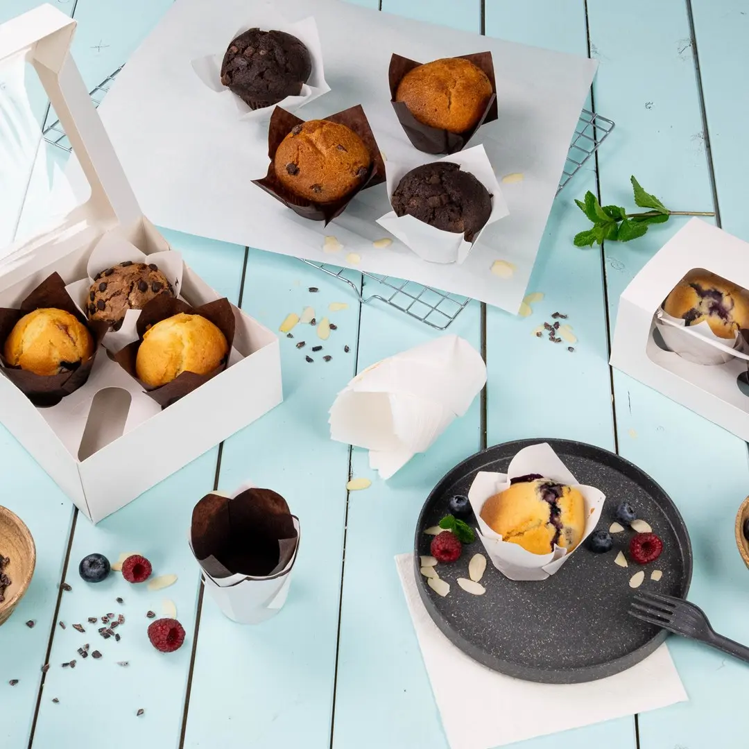 Assorted muffins in paper wrappers on a light blue surface, with a white box and a plate of muffins and berries.