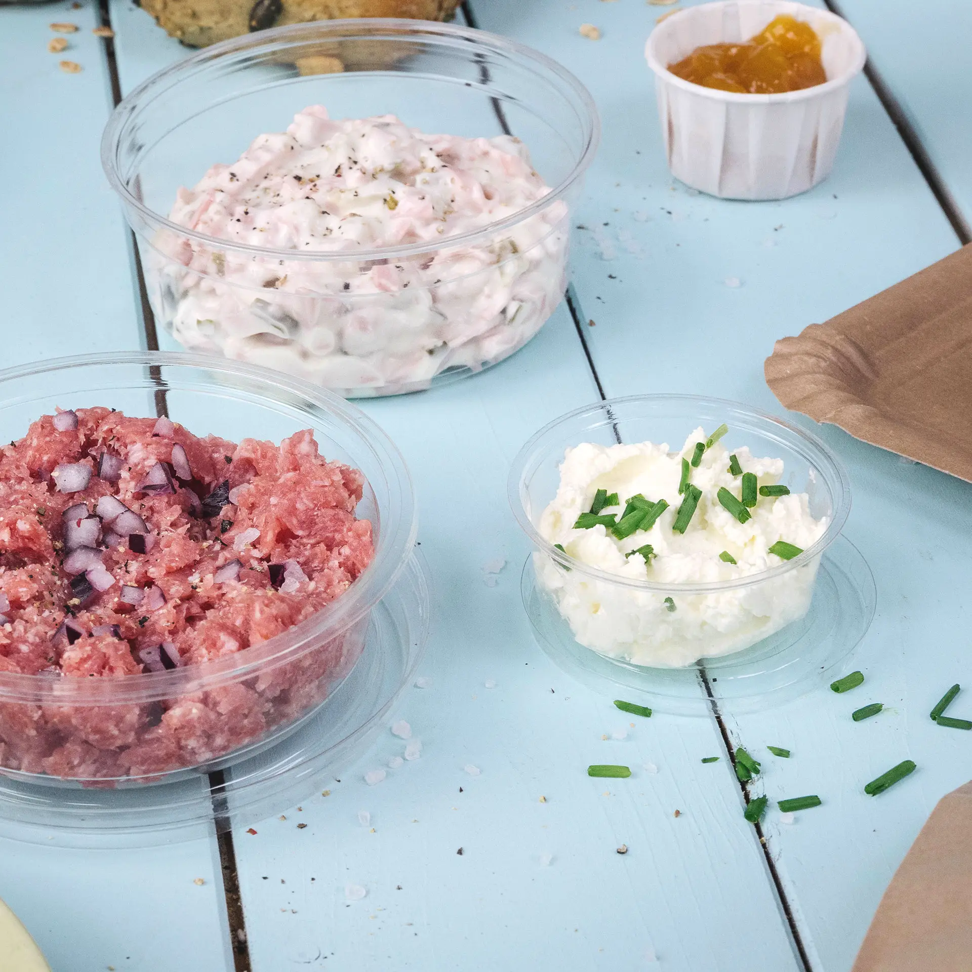 Drei transparente Portionsbecher mit verschiedenen Lebensmitteln auf einer hellblauen Holzoberfläche. Die Becher enthalten Hackfleisch, weißen Dip mit Schnittlauch und eine Sauce in einem kleinen Becher.