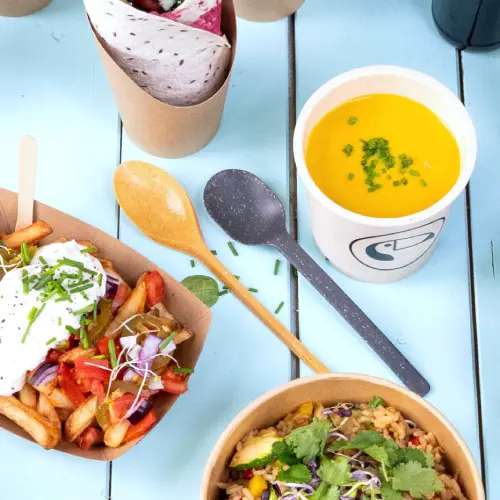 Bild zeigt verschiedene Lebensmittel wie Pommes mit Belag, eine Reisschüssel mit Kräutern, eine Suppe in einem Becher und wiederverwendbares Besteck auf einer blauen Oberfläche.