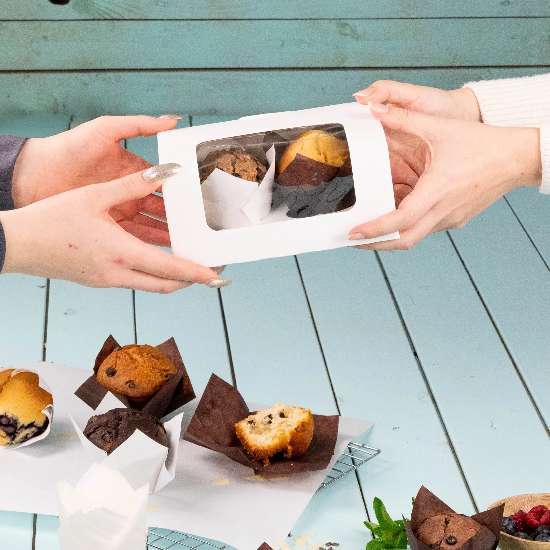 Weißer zweigeteilter Cupcake-Behälter mit Zellulosefenster, der übergeben wird und zwei Muffins enthält, auf einem hellblauen Tisch.