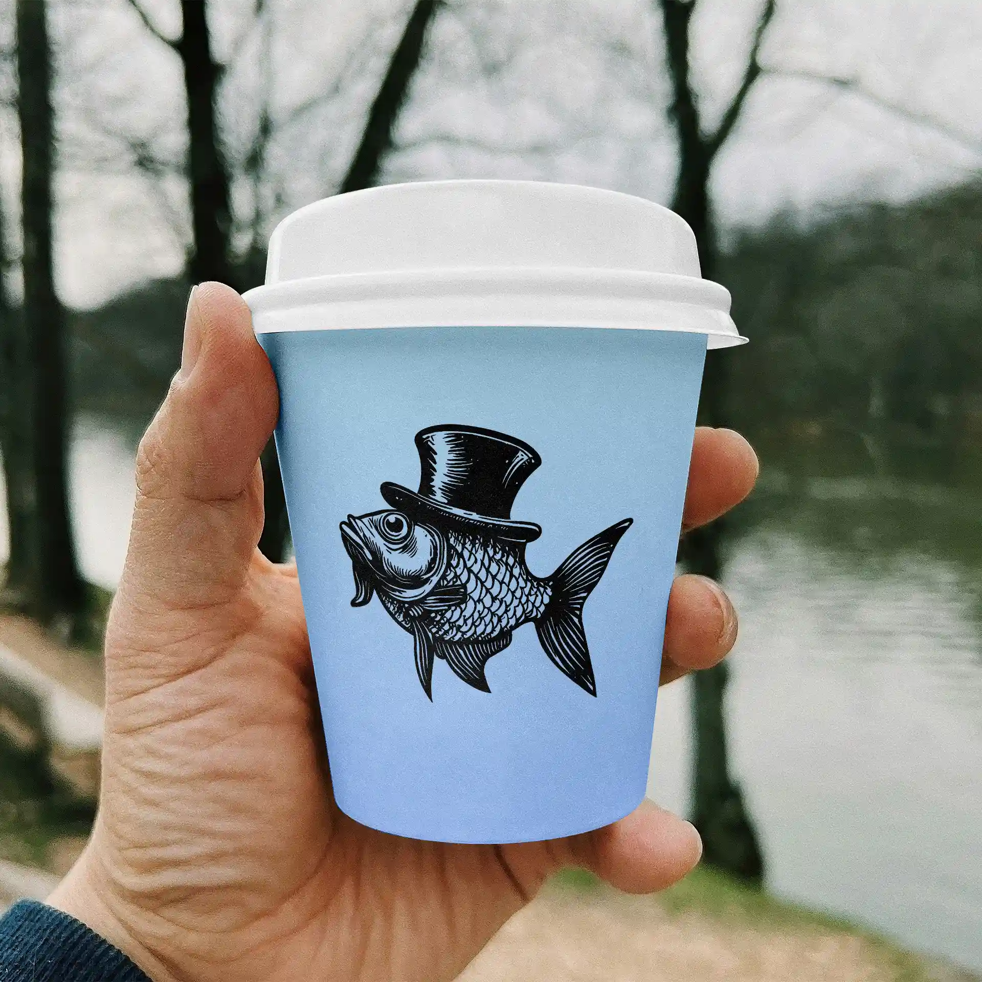 Hand holding a light blue paper cup featuring a black fish illustration wearing a top hat, with a blurred natural background.