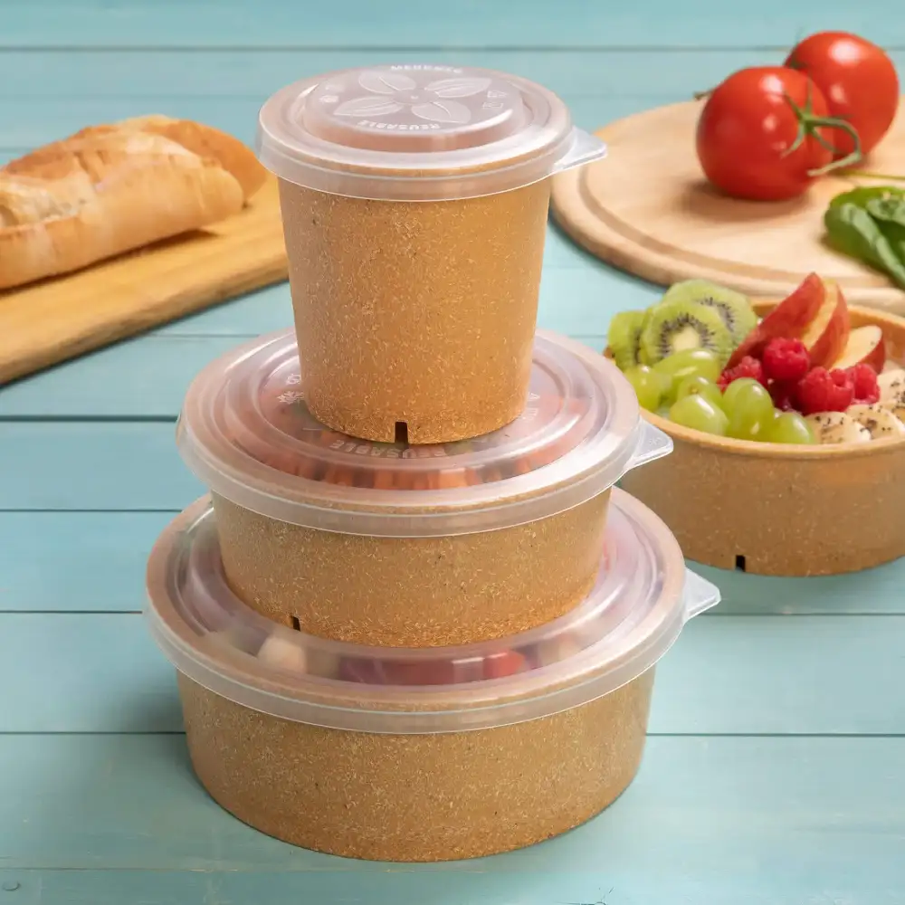 Stapel aus drei braunen Behältern mit klaren Deckeln auf einer blauen Fläche, mit Brot, Tomaten und Obstsalat in der Nähe.