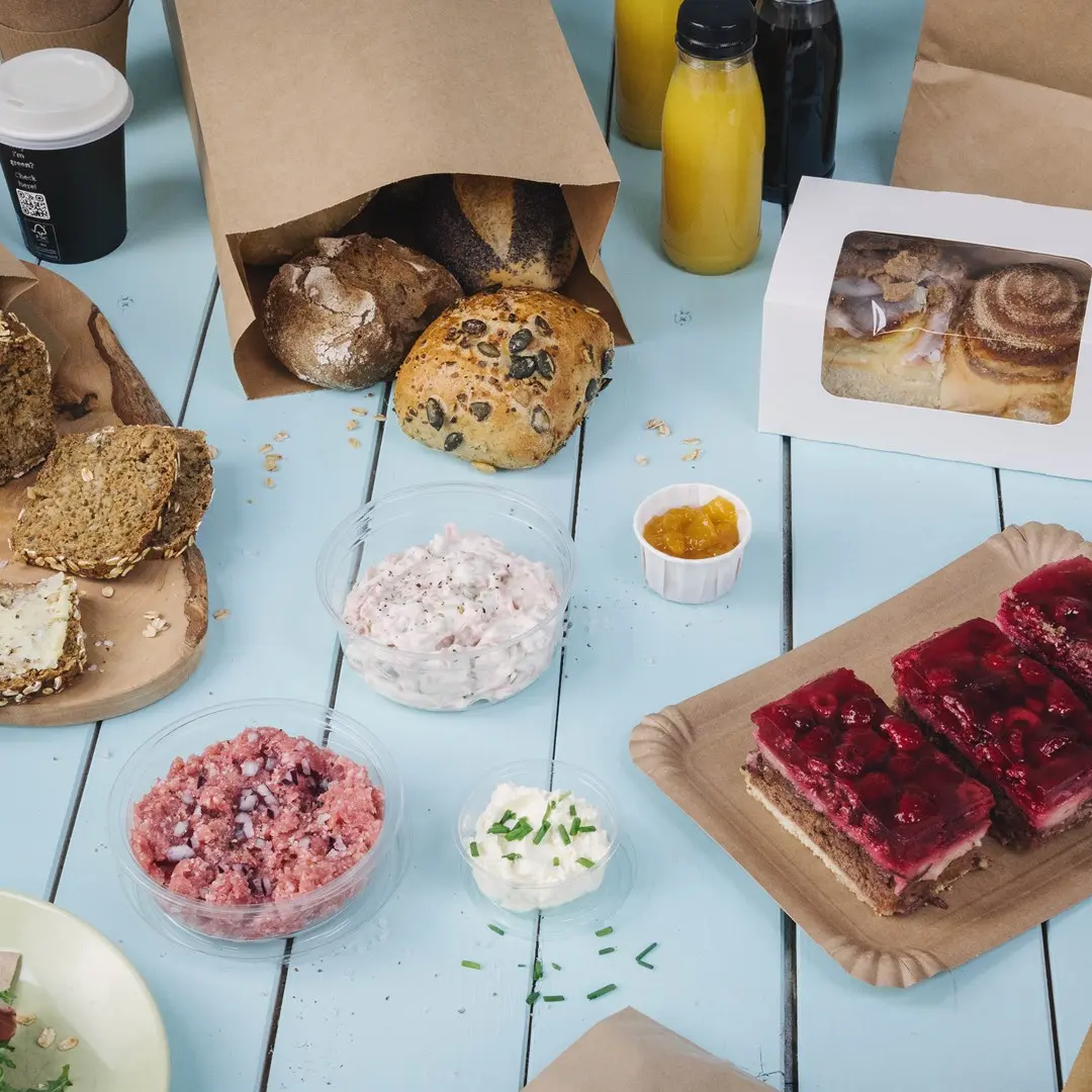 Verschiedene Backwaren, darunter Brötchen, Aufstriche, Desserts und Getränke auf einem blauen Holztisch.