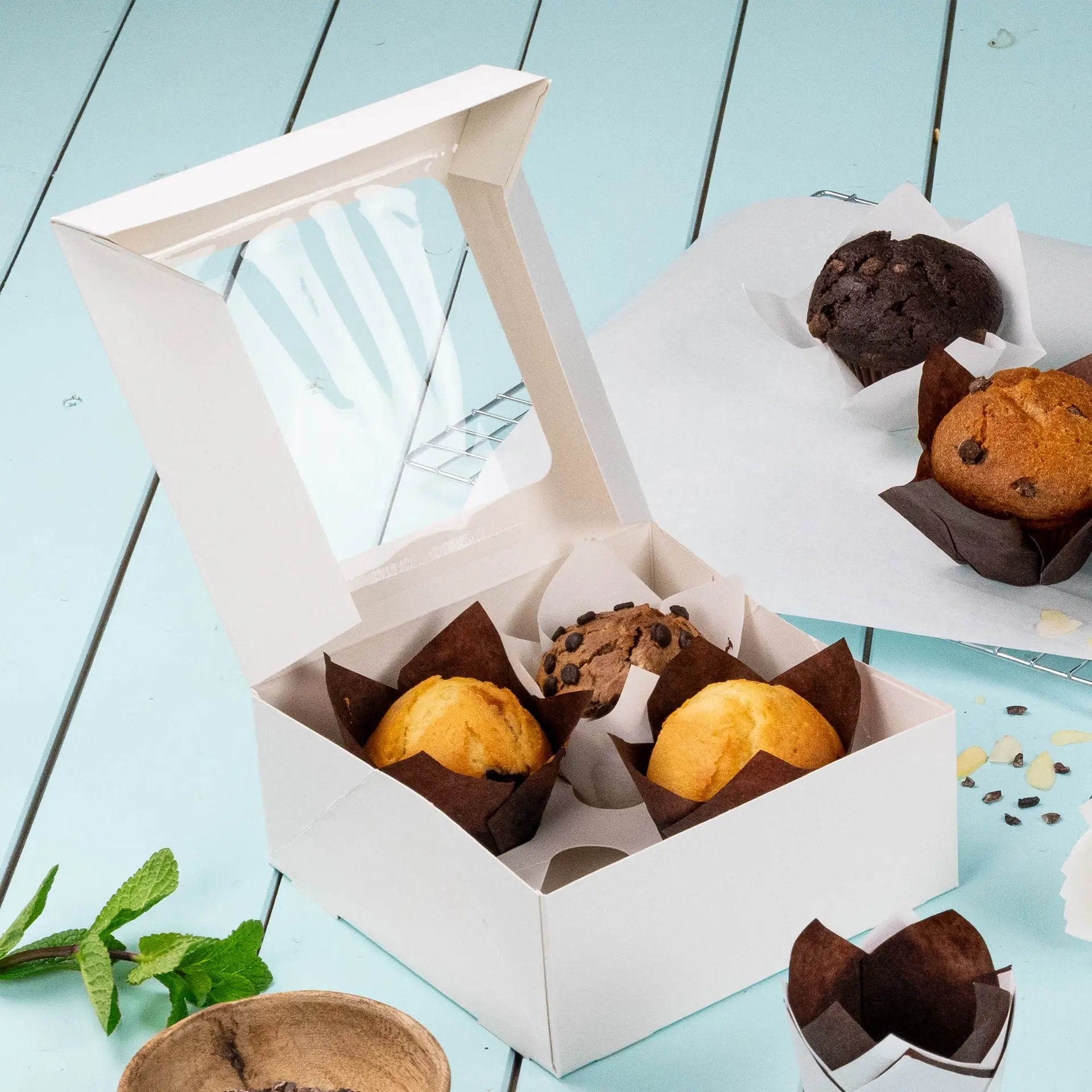 Weiße 4-teilige Cupcake-Box mit einer Cellulose- seitlichen Fenster, enthält Muffins und einen Schokoladenmuffin auf einer hellblauen Oberfläche.