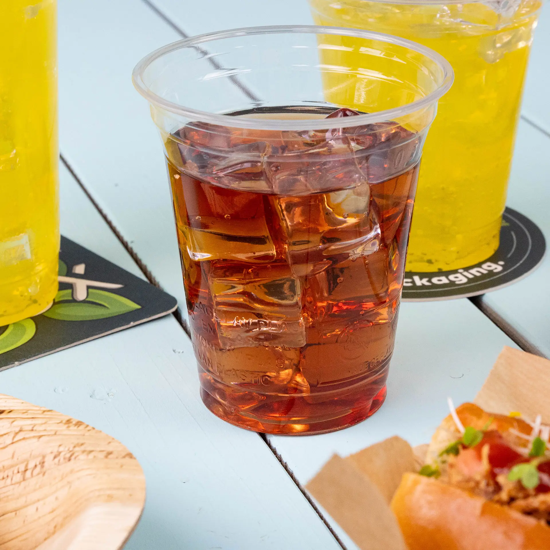 Transparenter 12 oz Plastikbecher aus PLA-Bioplastik mit Eistee, umgeben von gelben Getränken und einem Lebensmittel.